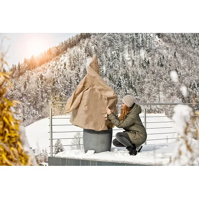 Windhager Winterschutzvlies - L x B: 5 x 2 m, Beige, Grammatur: 30 g/m² - Frau befestigt eine beigefarbene Pflanzenschutzhülle auf einem großen Topf auf einem verschneiten Balkon.