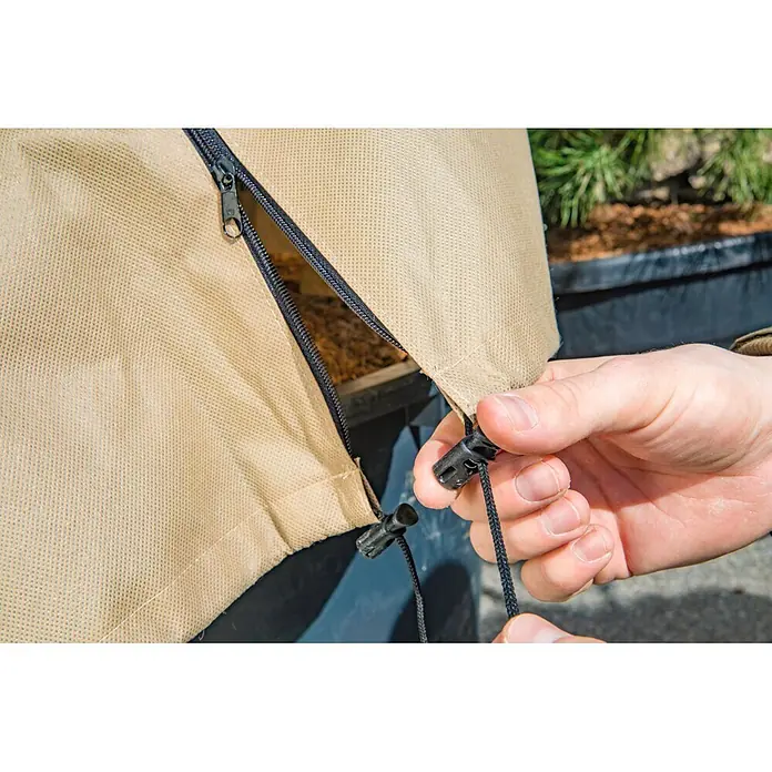Windhager Winterschutzvlies - L x B: 2,4 x 1,9 m, Beige, 140 g/m² - Eine Hand verstellt den Kordelzug einer hellbraunen Schutzhülle aus Stoff mit schwarzem Reißverschluss, die teilweise Mulch im Inneren zeigt.
