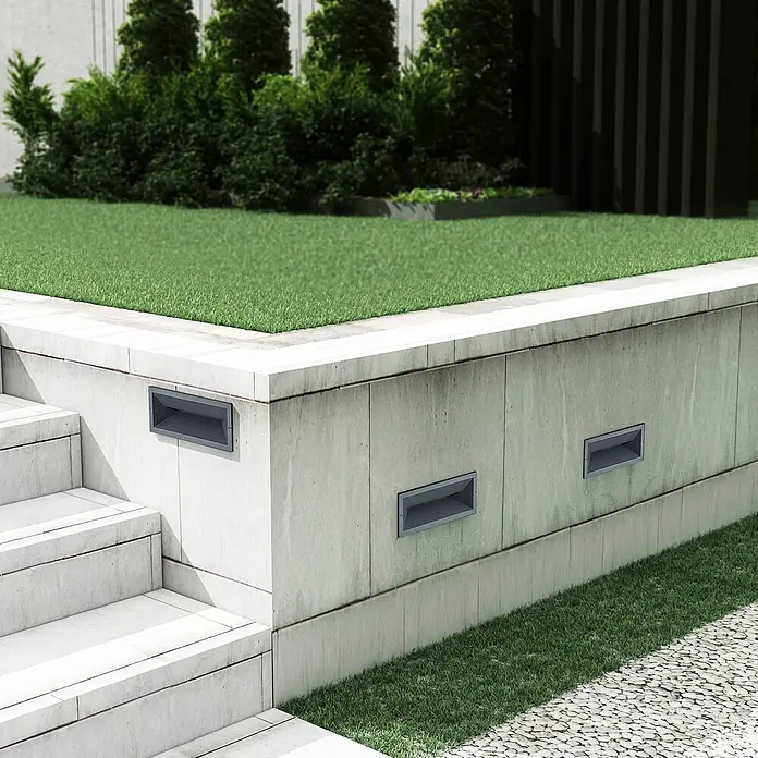 Lucande Außenwandleuchte Jaano - schwarz, IP65 - Rechteckige graue Einbau-Treppenleuchten in einer hellen Beton-Gartenmauer neben Stufen und Gras.