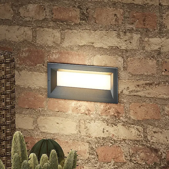 Lucande Außenwandleuchte Lachlain - alu / grau / zink, IP65 - Rechteckige anthrazitfarbene LED-Wandleuchte, beleuchtet an einer rustikalen Ziegelwand.