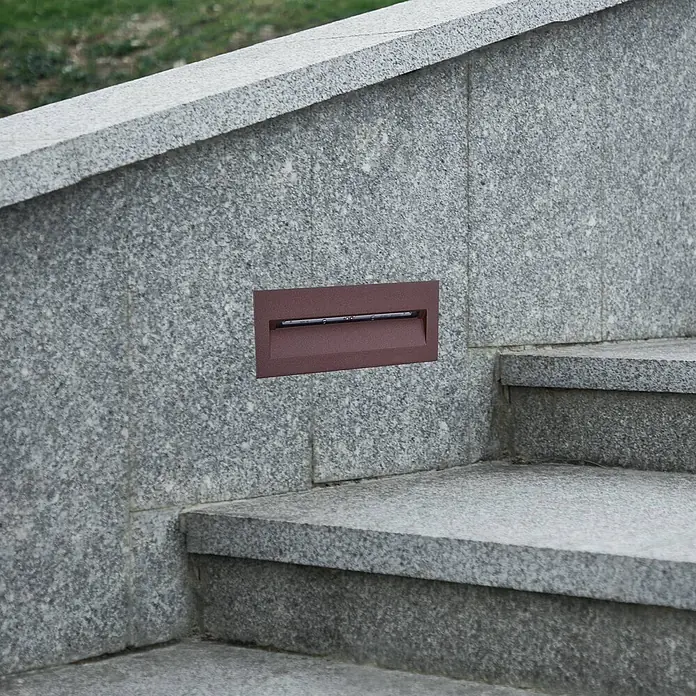 Lucande Außenwandleuchte Loya - braun / rost, IP54 - Einbaustufenleuchte in dunklem Bronze-Finish, in eine graue Granitwand neben Treppen integriert.