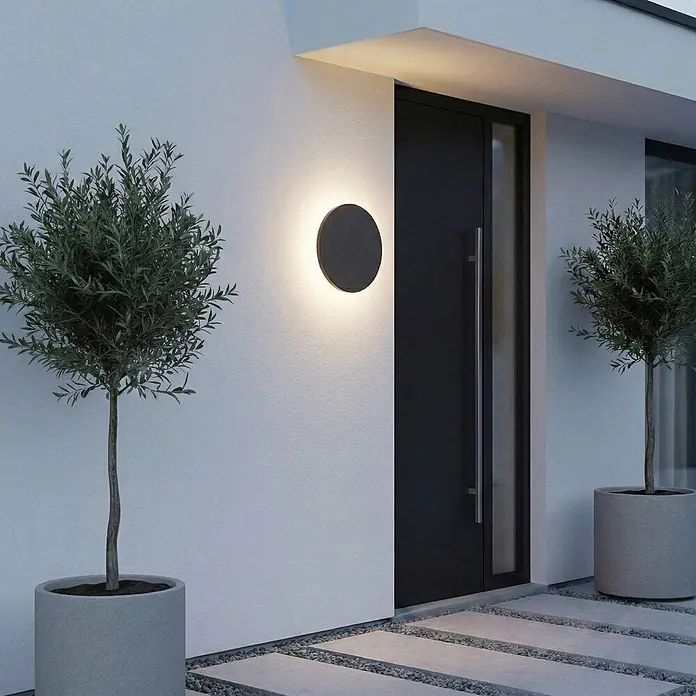 Lindby Außenwandleuchte Selinel - 5,3 x 23 x 23 cm, schwarz, schwarz, IP65 - Rundes, dunkles Wandlicht beleuchtet den Eingang eines modernen Hauses, montiert neben einer schwarzen Tür.