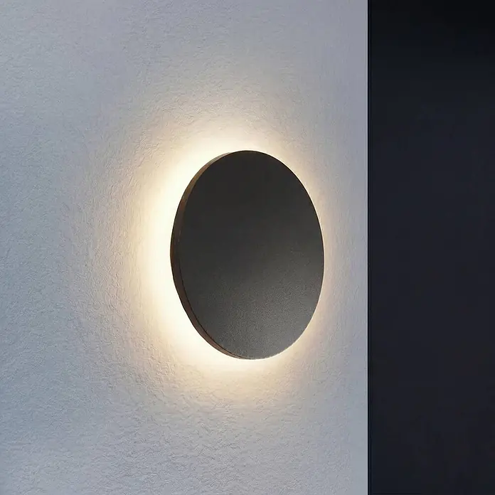 Lindby Außenwandleuchte Selinel - 5,3 x 23 x 23 cm, schwarz, schwarz, IP65 - Dunkelgraue runde Wandleuchte, die ein warmes, indirektes Licht auf eine texturierte Wand wirft.