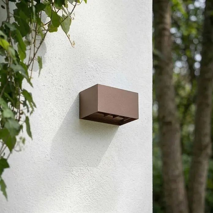 Lindby Außenwandleuchte Nivar - 10 x 20 x 10 cm, braun / rost, braun / rost, IP54 - Rechteckige bronzefarbene Außenwandleuchte an einer weißen texturierten Wand mit Efeu.