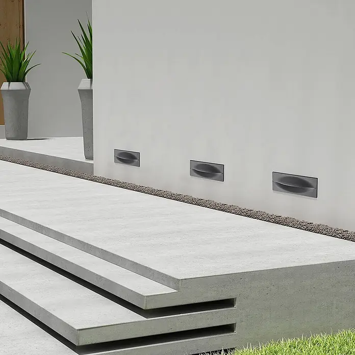 Lucande Außenwandleuchte Zandro - schwarz, IP65 - Drei dunkelgraue, eingelassene Außenwandleuchten mit geschwungenem Design, die einen Betonweg beleuchten.