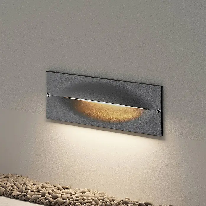 Lucande Außenwandleuchte Zandro - schwarz, IP65 - Rechteckige, dunkelgraue Außenstufenleuchte strahlt warmes Licht auf Wand und Kiesel.