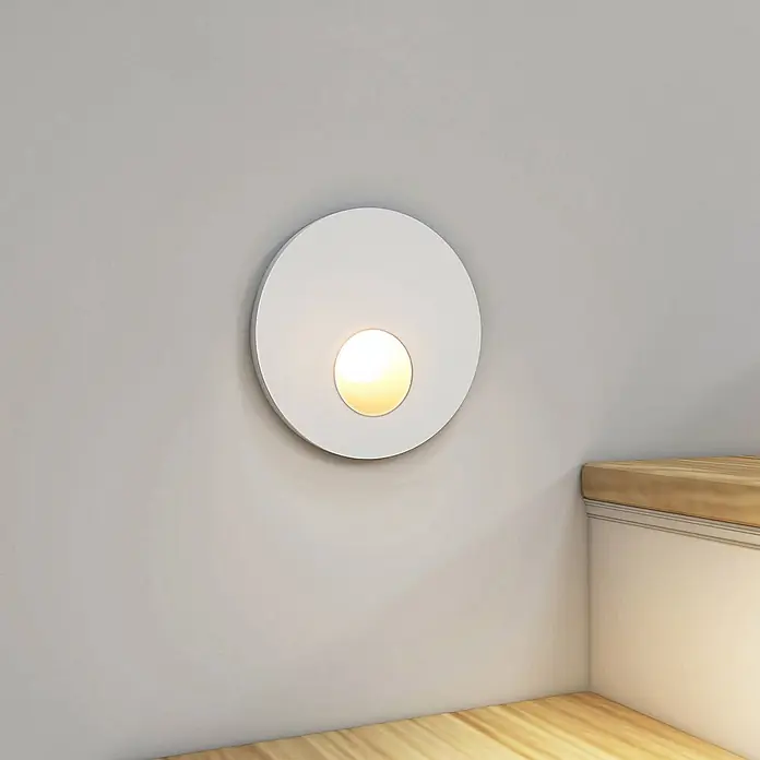 Arcchio Wandleuchte Vexi - L x B x H: 7,5 x 7,5 x 4,06 cm, weiß / opal - Weiße runde Wandleuchte mit warmem Lichtschein, an einer hellgrauen Wand über einer Holzecke montiert.