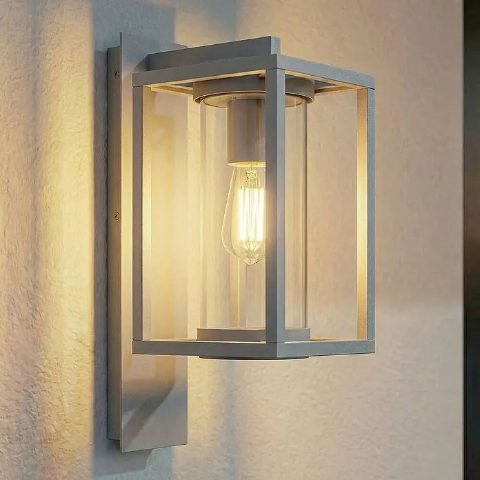 Lucande Außenwandleuchte Ferda - alu / grau / zink, alu / grau / zink, IP44 - Moderne graue Außenwandleuchte mit Glaszylinder und beleuchteter Edison-LED-Birne an strukturierter Wand.