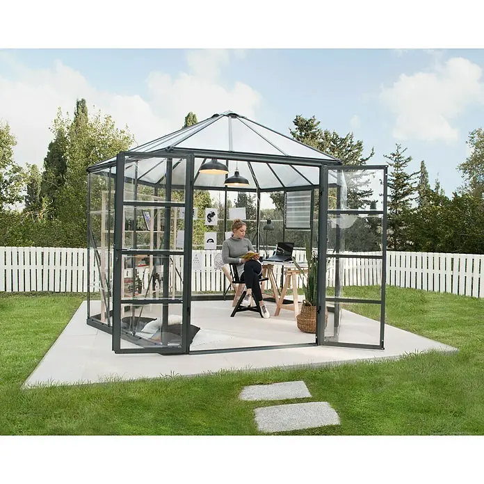 Palram – Canopia Invernadero Oasis - Medidas ext. (An x Pr): 316 x 362,8 cm, 0,7 mm, Antracita - Una mujer sentada en un invernadero con un escritorio y una estantería leyendo un libro. El invernadero está situado en un patio en el jardín.
