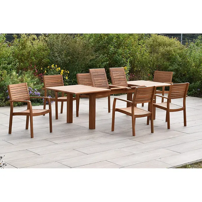 Merxx Gartentisch-Set Batavia - Gesamtumfang: 7 -tlg., L x B x H: 260 x 100 x 75 cm, Akazie, Stapelbar - Gartensitzgruppe aus Holz auf einer Terrasse, umgeben von Pflanzen. Ausziehbarer Tisch und Stühle aus hellbraunem Holz.