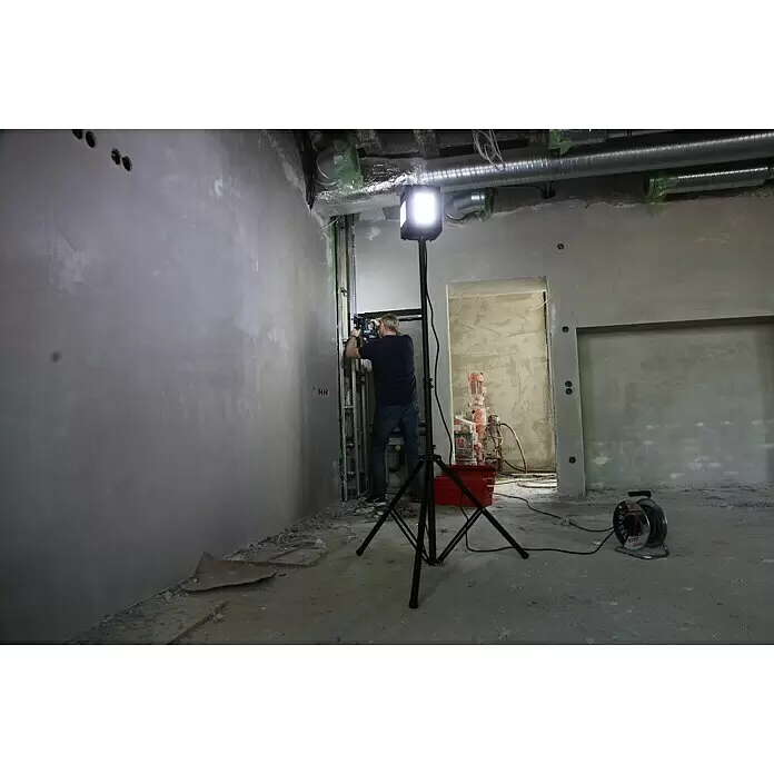 Profi Depot Proyector LED 2 cabezales - 50 W, 8.000 lm, 6.000 K - 7.000 K, L x An x Al: 1,14 x 1,32 x 2,25 m, IP54 - Foco de trabajo negro sobre trípode, iluminando a un obrero en una obra en construcción.