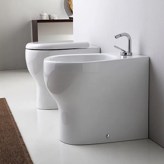 Kiamami Valentina Stand-WC-Kombination - Mit Spülrand, WC Abgang: Senkrecht, Weiß - Zwei weiße Keramik-Badezimmerarmaturen: eine Toilette mit geschlossenem Deckel und ein Bidet mit Chromarmatur, nebeneinander platziert.
