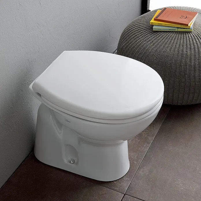 Kiamami Valentina Stand-WC-Kombination - Mit Spülrand, WC Abgang: Senkrecht, Weiß - Weißes Keramik-WC mit geschlossenem Deckel auf dunklen Fliesen, neben einem grauen Strickpouf.