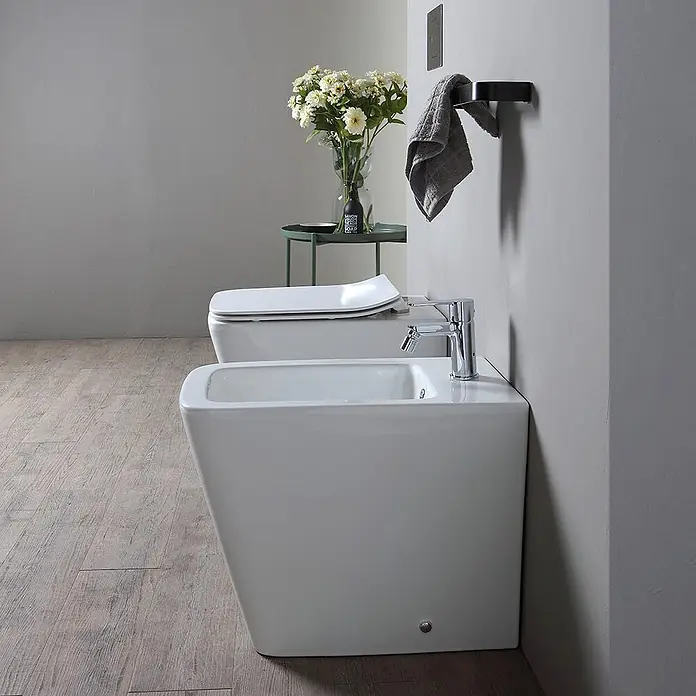 Kiamami Valentina Stand-WC-Kombination - Spülrandlos, WC Abgang: Senkrecht, Weiß - Ein modernes weißes Keramik-Bidet und passendes WC in einem minimalistischen Badezimmer mit Holzfußboden.