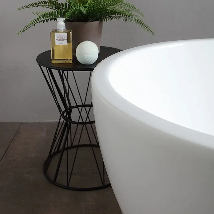 Kiamami Valentina Freistehende Badewanne - L x B: 1.700 x 800 cm, ABS-Kunststoff und Acryl, Weiß - Elegante weiße Badewanne in einem Badezimmer, daneben ein schwarzer Beistelltisch mit Badeutensilien und Pflanze.