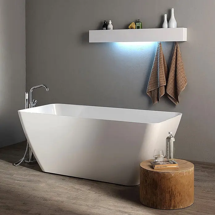 Kiamami Valentina Freistehende Badewanne - L x B: 1.590 x 705 cm, ABS-Kunststoff und Acryl, Weiß - Weiße, moderne freistehende Badewanne in einem Badezimmer mit Bodenarmatur und einem Holztischchen mit Wasserflasche und Buch.