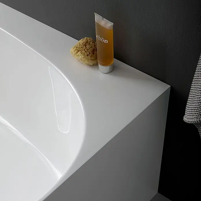 Kiamami Valentina Freistehende Badewanne - L x B: 1.580 x 860 cm, ABS-Kunststoff und Acryl, Weiß - Weiße, glänzende Badewanne mit Naturschwamm und Badegel am Rand vor dunkler Wand.