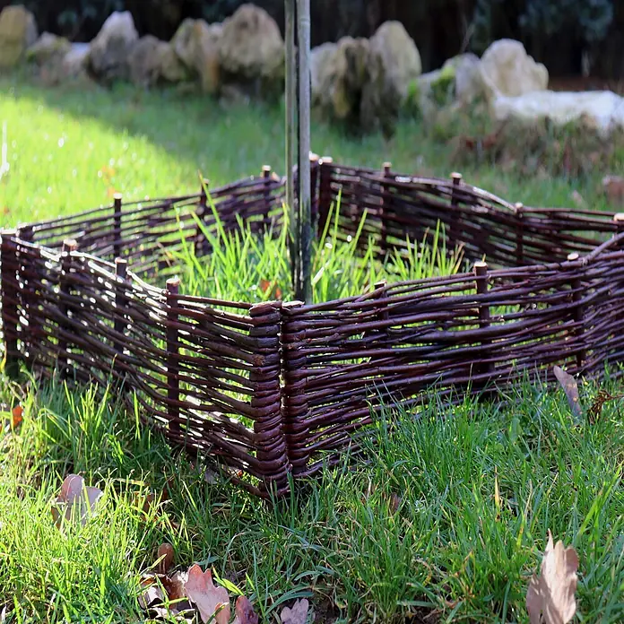 Aquagart Steckzaun-Element 8 30x60cm Beeteinfassungen aus Weide I - braun, Holz - Ein dunkelbrauner Weidenflechtzaun umgibt die Basis einer Pflanze in einem grünen Garten.