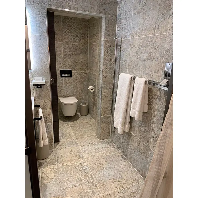 Feinsteinzeugfliese Carpet Natural - 59,2 x 59,2 cm, Sand, Matt - Modernes Badezimmer mit aufwendigen grau-beigen Musterfliesen, einer weißen Wandtoilette und weichen weißen Handtüchern.