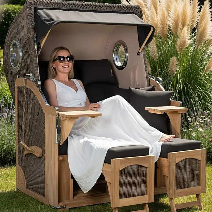 Mr. Deko Strandstuhl Strandkorb Düne Bullauge Teak PE Grey Dessin 548 - 1 Stk., Teakholz. Polyrattan, Rückenlehne verstellbar, PE Grey - Luxuriöser Strandkorb aus braunem Korbgeflecht und Holz mit dunklen Kissen und beigem Verdeck, in dem eine Frau bequem im Garten entspannt.