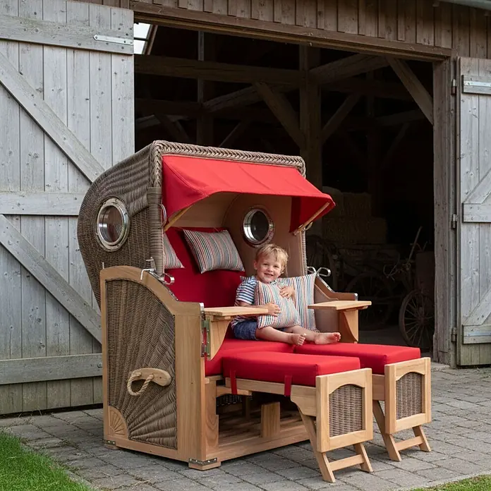Mr. Deko Strandstuhl Strandkorb Düne Bullauge Teak PE Grey Dessin 585 - 1 Stk., Teakholz. Polyrattan, Rückenlehne verstellbar, PE Grey - Ein Kind sitzt lächelnd in einem Strandkorb aus Korbgeflecht und Holz mit roten Polstern und Verdeck.