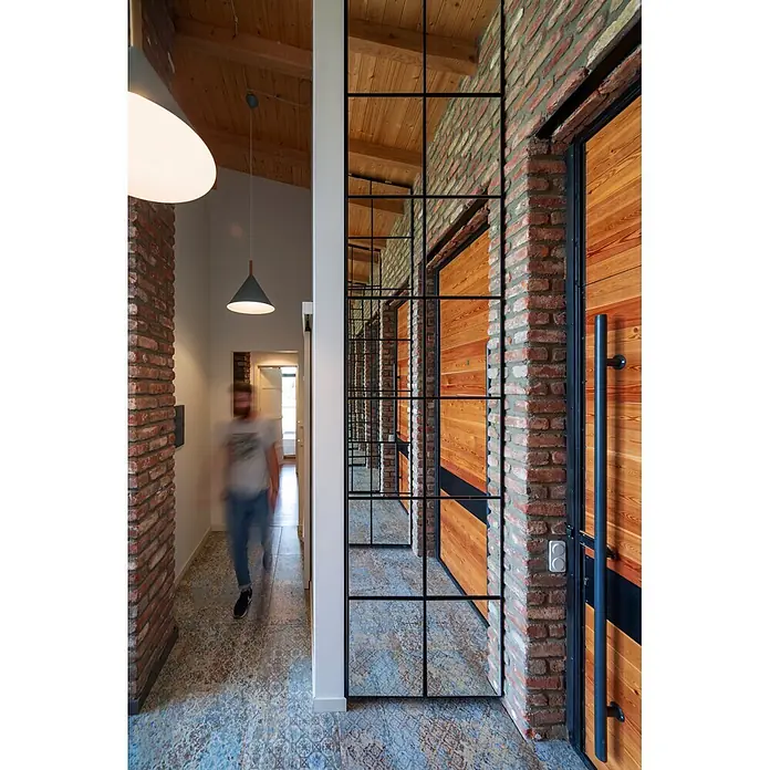 Feinsteinzeugfliese Carpet Natural - 59,2 x 59,2 cm, Vestige, Matt - Flur im Industriestil mit schwarzgerahmter Spiegelwand, Ziegelwänden und Holzdecke. Eine Person geht über gemusterte Fliesen.
