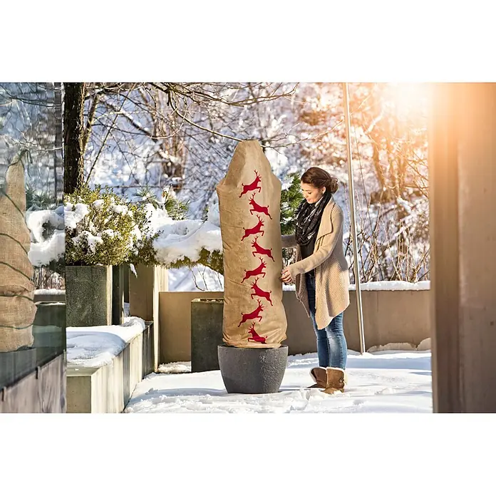 Windhager Winterschutzhaube Protect - L x B: 1,8 x 0,6 m, Motiv: Hirsch - Frau justiert hohen beigen Pflanzenschutz mit roten Rentier-Designs im verschneiten Garten.