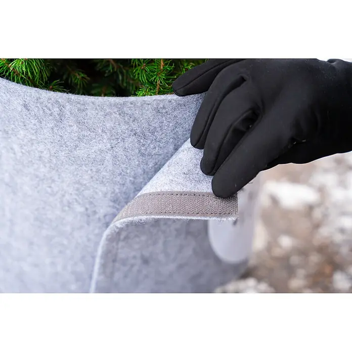 Windhager Dekoartikel Topfschutz Hello Winter - Grau, Frostschutz, Länge: 25 cm, Rund - Eine behandschuhte Hand richtet eine graue Filz-Winterschutzhülle um eine grüne Pflanze, ein Befestigungsband ist sichtbar.