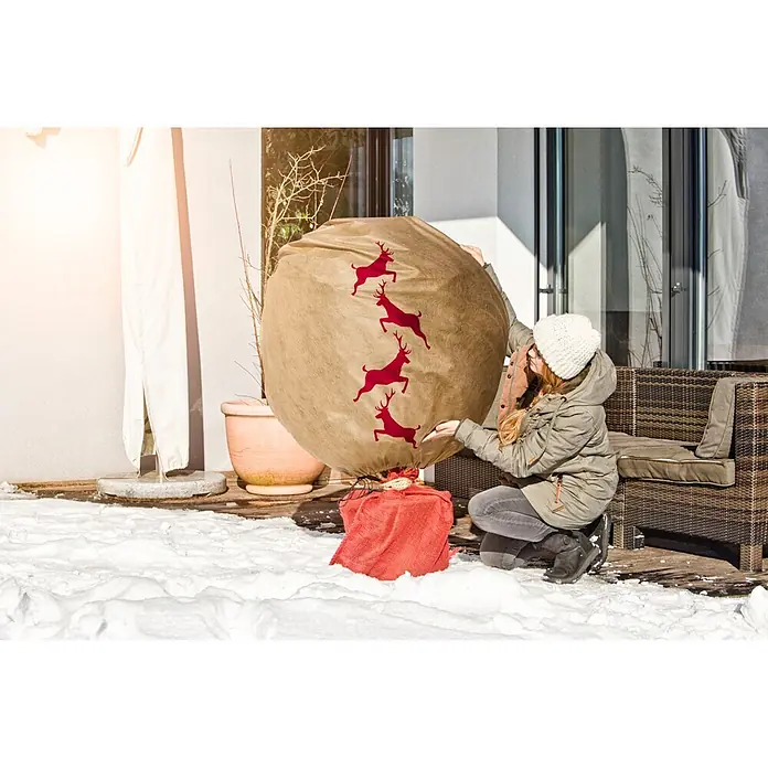 Windhager Winterschutzhaube Protect - L x B: 1,1 x 1,1 m, Motiv: Hirsch - Großer khakifarbener Sack mit roten Hirschsilhouetten, unten gebunden, von einer Person in verschneiter Außenumgebung gehandhabt.