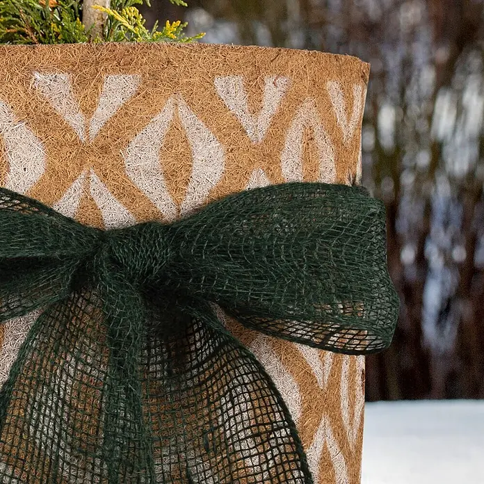 Windhager Winterschutz Kokos-Filzmatte Oriental - L x B: 150 x 50 cm - Runder Pflanzkübel aus Naturfaser mit weißem geometrischem Muster, verziert mit einer dunkelgrünen Netzwirkschleife. Oben ist Pflanzengrün sichtbar.
