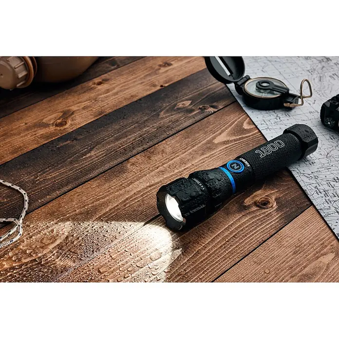 Nebo Tools Linterna Blueline 1800 - 16 x 3 x 3 cm, 180 lm - 1.800 lm, IPX7 - Linterna negra y húmeda sobre una superficie de madera, iluminando gotas de agua. Una brújula y un mapa están cerca.