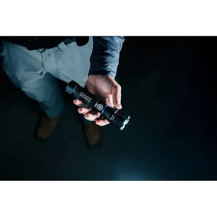 Nebo Tools Linterna Blueline 1800 - 16 x 3 x 3 cm, 180 lm - 1.800 lm, IPX7 - Una mano sostiene una linterna negra con detalles azules, emitiendo luz brillante en un entorno tenue.