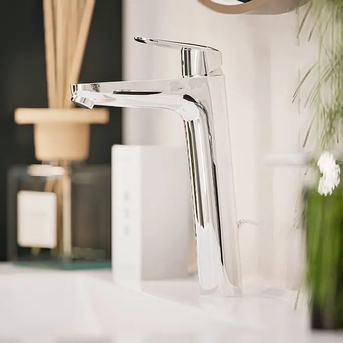 VitrA Flow Round Waschtischarmatur - Höhe: 27 cm, Ausladung Armatur: 151 mm, Chrom, Mit Zugstange - Polierte Chromarmatur auf einem weißen Waschbecken, mit verschwommener Badezimmerdekoration im Hintergrund.