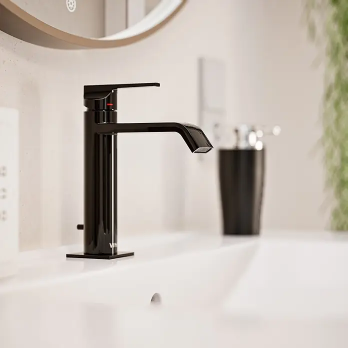 VitrA Suit Waschtischarmatur - Schwarz, Glänzend, Höhe: 19 cm - Schwarze moderne Armatur auf einem weißen Waschbecken, mit Seifenspender und unscharfer Pflanze im Hintergrund.