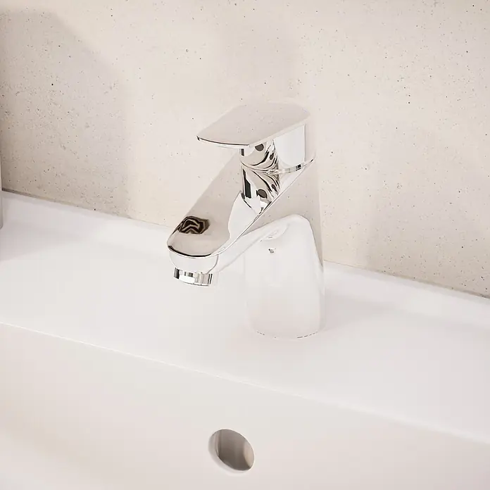 VitrA Flow Round Waschtischarmatur - Höhe: 15 cm, Ausladung Armatur: 114 mm, Chrom, Ohne Zugstange - Verchromte Armatur mit weißem Sockel auf einem weißen Waschbecken vor einer hellen, strukturierten Wand.