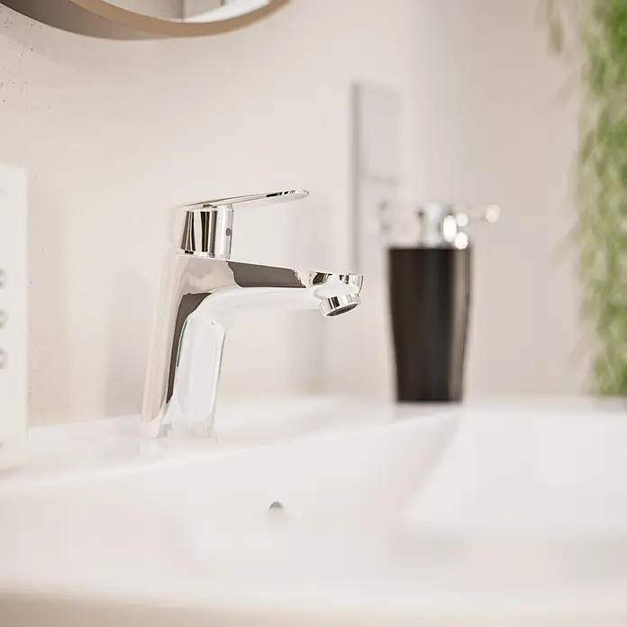 VitrA Flow Round Waschtischarmatur - Höhe: 15 cm, Ausladung Armatur: 114 mm, Chrom, Ohne Zugstange - Moderner Chrom-Wasserhahn in einem weißen Waschbecken. Ein schwarzer Seifenspender ist im unscharfen Hintergrund.