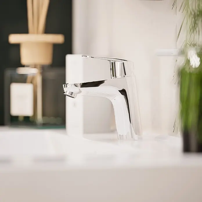 VitrA Flow Round Waschtischarmatur - Höhe: 15 cm, Ausladung Armatur: 114 mm, Chrom, Ohne Zugstange - Ein moderner, polierter Chrom-Wasserhahn auf einem weißen Waschbecken, mit unscharfen Dekoartikeln im Hintergrund.
