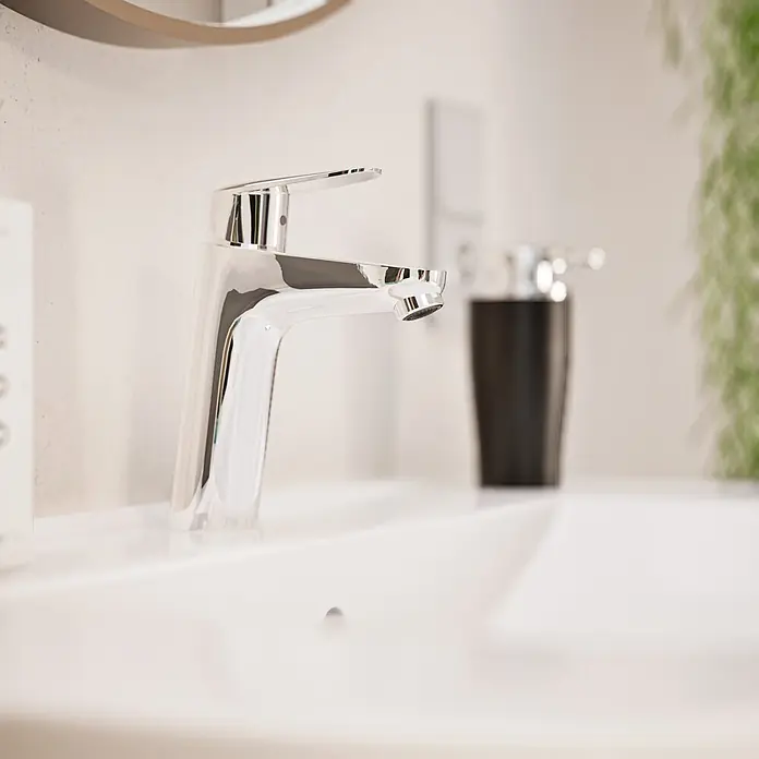 VitrA Flow Round Waschtischarmatur - Höhe: 18 cm, Ausladung Armatur: 115 mm, Chrom, Ohne Zugstange - Glänzende Chromarmatur in einem weißen Waschbecken, mit einem Seifenspender im unscharfen Hintergrund.
