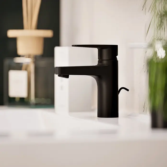 VitrA Flow Square Waschtischarmatur - Schwarz, Matt, Höhe: 15,6 cm, Mit Zugstange - Mattschwarzer Badarmatur in einem weißen Waschbecken, mit verschwommenen Dekoelementen im Hintergrund.