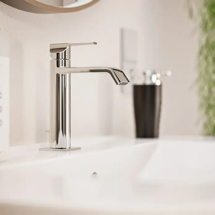 VitrA Suit Waschtischarmatur - Chrom, Glänzend, Höhe: 19 cm - Ein schlanker, moderner Chromwasserhahn auf einem weißen Waschbecken, mit unscharfem Seifenspender und Pflanze im Hintergrund.