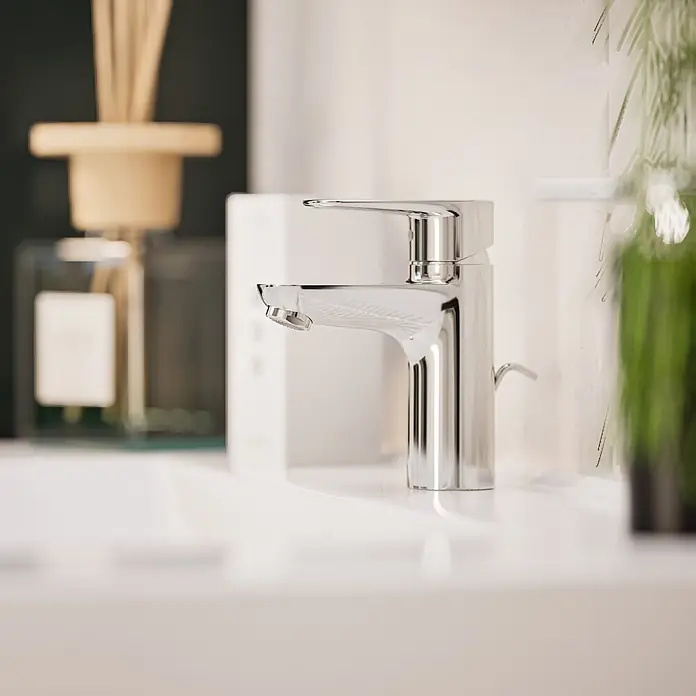 VitrA Flow Soft Waschtischarmatur - Chrom, Höhe: 15 cm, Mit Zugstange - Verchromte Armatur auf einem weißen Waschbecken im Badezimmer, mit unscharfen Badartikeln im Hintergrund.