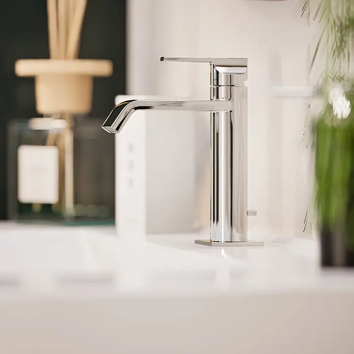 VitrA Suit Waschtischarmatur - Chrom, Glänzend, Höhe: 19 cm - Moderner Chrom-Badarmatur auf einem weißen Waschbecken, mit unscharfen Badaccessoires im Hintergrund.
