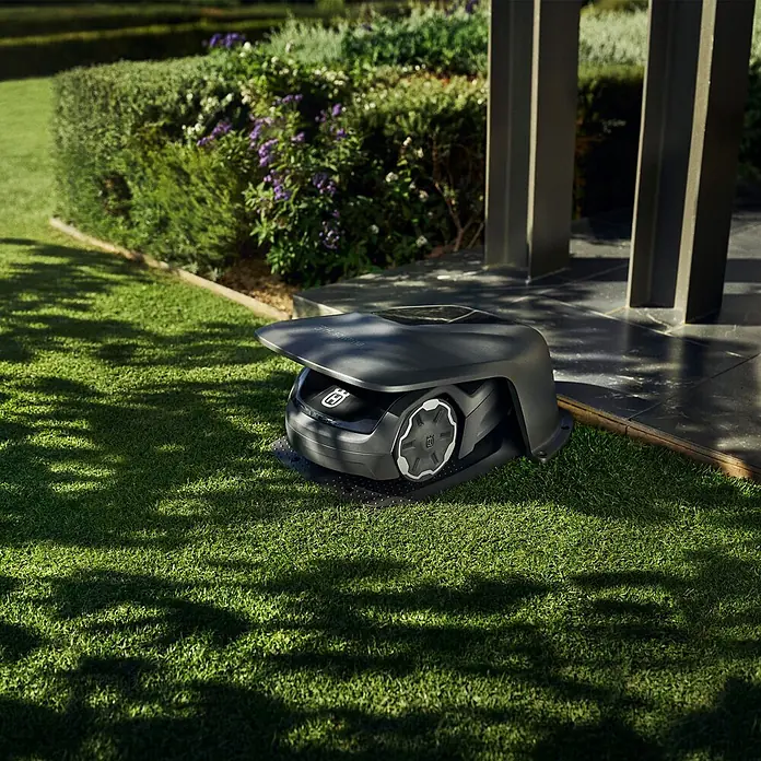 Husqvarna Mähroboter-Garage Aspire R4 - Grau, Aspire R4 - Dunkelgrauer Mähroboter steht auf Rasen neben einer Terrasse vor einer Gartenhecke.