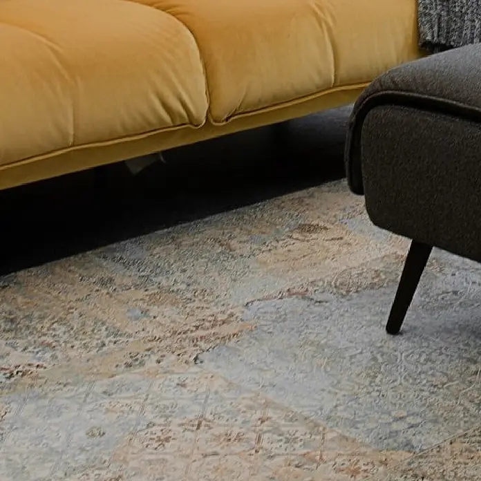 Feinsteinzeugfliese Carpet Natural - 59,2 x 59,2 cm, Vestige, Matt - Distressed Teppich in Blau-, Grau- und Rosttönen, teilweise unter gelbem Sofa und neben grauem Ottoman.