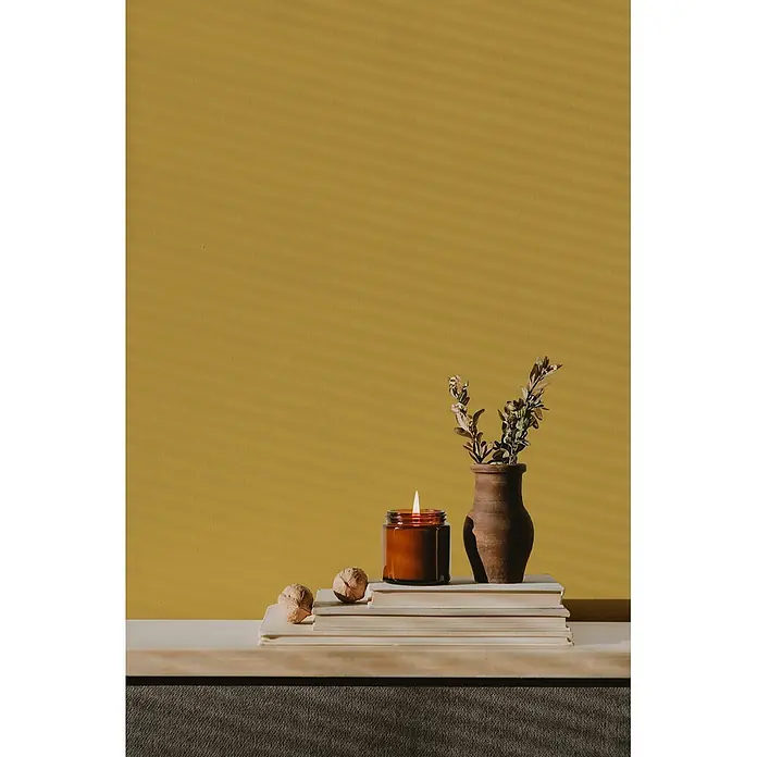 Livingwalls Wandfarbe Gelb Sunny Yellow DD126102 5l - Gelb, Matt, 5 l - Eine brennende Kerze in bernsteinfarbenem Glas, eine rustikale Vase mit Zweigen und Walnüsse auf gestapelten Büchern auf Holz.