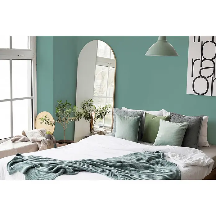 Livingwalls Wandfarbe Grün Emerald Green DD125944 1l - Grün, Matt, 1 l - Ein ruhiges Schlafzimmer mit einem Bett mit weißer Bettwäsche, weichen grünen und grauen Kissen und einer gestrickten tealfarbenen Decke. Ein Bogenspiegel und Pflanzen tragen zur ruhigen Atmosphäre bei.