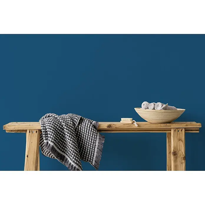Livingwalls Wandfarbe Blau Classic Cornflower 3005 2,5l - Matt, 2,5 l - Holzbank mit gemusterter grauer Decke, einer Holzschale mit gerollten grauen Handtüchern und einer kleinen Bürste.