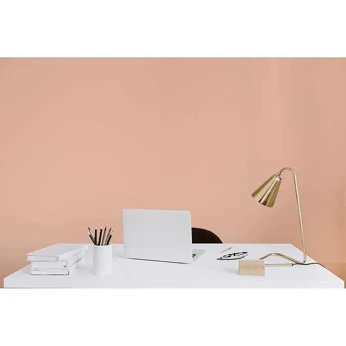 Livingwalls Wandfarbe Rosa Honeydew Melon 6022 2,5l - Matt, 2,5 l - Ein minimalistischer weißer Schreibtisch mit Laptop, Büchern und einer goldenen Lampe vor einer warmen pfirsichfarbenen Wand.