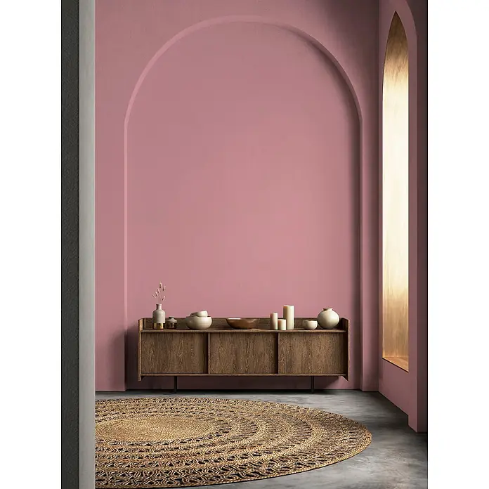Tadessi Wandfarbe Pfirsich - Pink, Matt, 2,5 l - Ein Holz-Sideboard und ein runder Jute-Teppich möblieren einen Raum mit rosa Bogenwand.