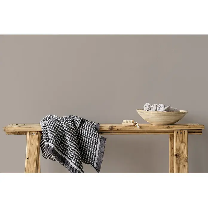Livingwalls Wandfarbe Taupe Whole Grain 1018 5l - Matt, 5 l - Rustikale Holzbank mit schwarz-weiß gemusterter Decke, Holzschale mit gerollten grauen Handtüchern und Holzbürste.